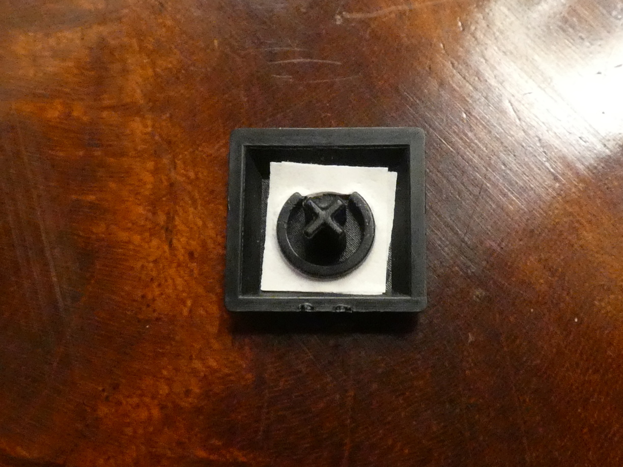 A close-up photo of one of the slightly smaller top row keycaps with the bottom side up. It features a thin open ring with a diameter of about half the key. Inside this ring is the x-shaped shaft which looks similar to those dog treats or something. Outside of the ring a stack of two paper squares with cut-out circles were placed to solve the top-row typing issue.