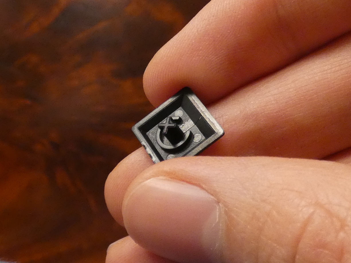 A close-up photo of one of the slightly smaller top row keycaps with the bottom side up held by my fingers that look giant in comparison. It features a thin open ring with a diameter of about half the key. Inside this ring is the x-shaped shaft which looks similar to those dog treats or something.