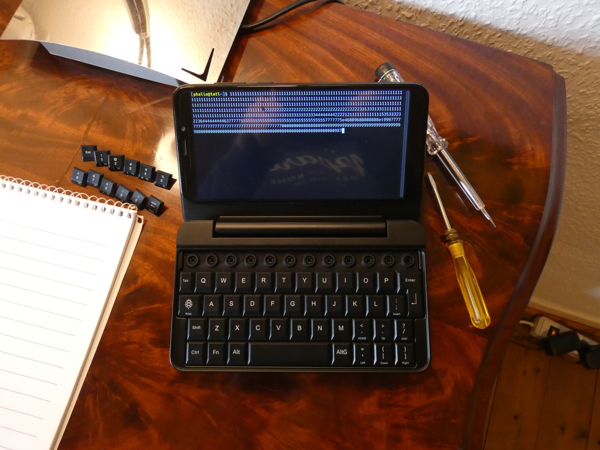 A photo of the PinePhone Keyboard on a table with the top row of keycaps removed, exposing the protruding membrane rings below. The removed keycaps are to the left of the device and two small screwdrivers are to its right.